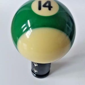 May include: Green and white billiard ball with the number 14 printed on it. The ball is attached to a black gear shift knob.