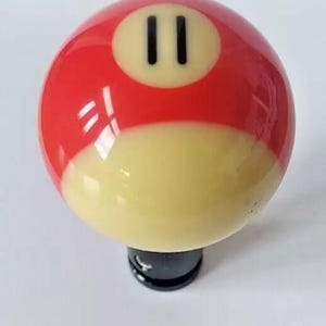 May include: A red and white billiard ball with the number "11" in black on it. The ball is attached to a black base.
