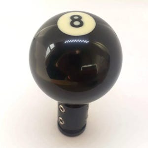 May include: Black and white 8-ball gear shift knob. The knob is made of a smooth, shiny material and has a black base with two holes.