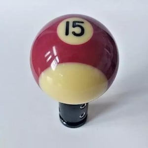 May include: A red and white billiard ball with the number 15 on it. The ball is attached to a black gear shift knob.