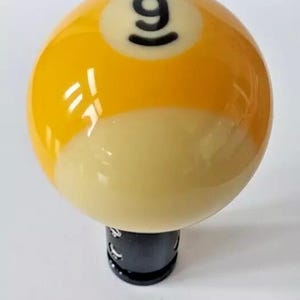 May include: A yellow and white billiard ball with the number 9 on it. The ball is mounted on a black base.