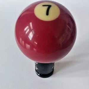 May include: A red billiard ball with a white number 7 on it. The ball is mounted on a black base.