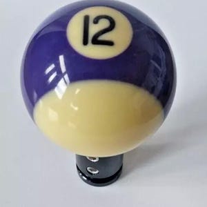 May include: A custom shift knob for a vehicle, featuring a purple and yellow billiard ball design. The ball is numbered with a black "12" on a yellow circle. The knob is mounted on a black base.