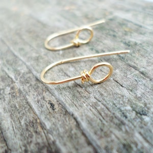 May include: A pair of gold-toned earrings with a simple knot design. The earrings are made of thin wire and have a delicate, minimalist look.