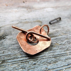 May include: A copper-colored metal pendant with a swirling pattern and a small loop at the top. The pendant is attached to a copper-colored metal wire that is bent into a loop.