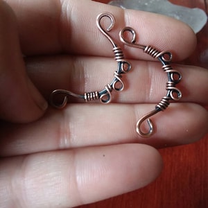 May include: Two handcrafted copper-colored wire earrings with a unique, curved design. Each earring features a series of loops and wrapped wire accents. The earrings are held in a hand, showcasing their intricate details and artistic craftsmanship.
