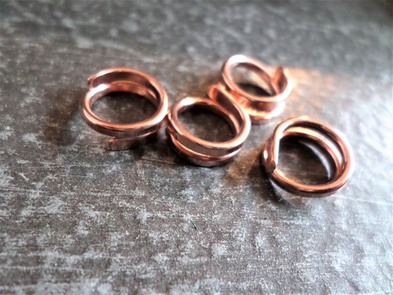K&ouml;nnte beinhalten: F&uuml;nf kupferfarbene Sprungringe, jeder mit einem Doppel-Loop-Design. Die Ringe sind in einer Gruppe auf einer strukturierten grauen Oberfl&auml;che angeordnet.