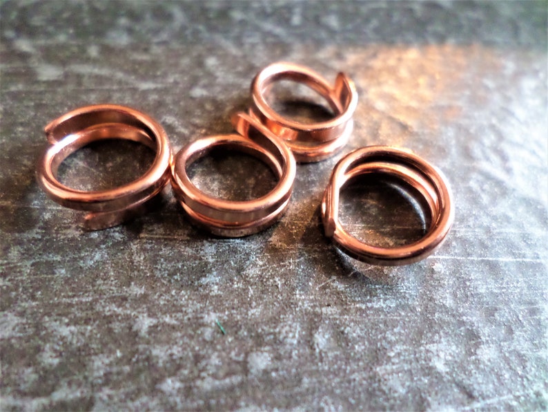 K&ouml;nnte beinhalten: Vier Kupferringe, jeder mit einem doppelt gewickelten Design, liegen auf einer strukturierten Oberfl&auml;che. Die Ringe haben eine warme, rotbraune Farbe.