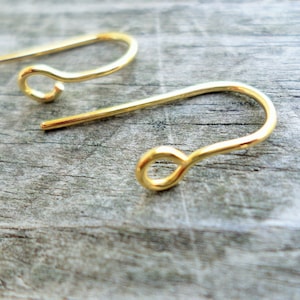 May include: Two gold-colored earring hooks, also known as ear wires, are shown on a wooden surface. The hooks are made of metal and have a simple, classic design.