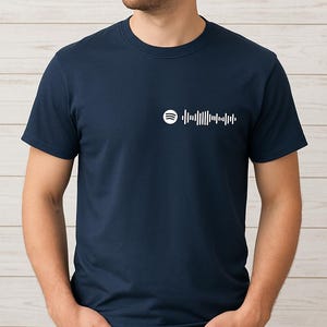 May include: Navy blue t-shirt featuring a white Spotify code design. The short-sleeved top is made of a soft material and is suitable for casual wear. The person in the image is wearing blue jeans.