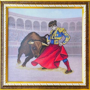 May include: Cross-stitch artwork depicting a bullfight scene. A matador in a blue and gold suit and red cape faces a charging brown bull in a bullring. The artwork is framed in a gold-colored frame.