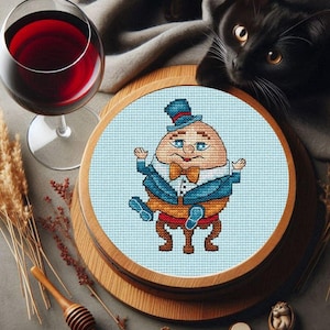 Op de afbeelding: Een kruissteekborduursel van Humpty Dumpty die op een muur zit, gekleed in een blauw pak, een strikje en een hoge hoed. De borduursel is ingelijst in een ronde houten ring.