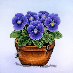 May include: Cross-stitch embroidery of a potted plant with purple pansies and green leaves. The terracotta pot has a small spider on the side. The embroidery is on a white background.