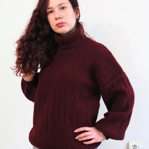Op de afbeelding: Een bordeauxrode trui met een hoge hals en een grove breistructuur. De trui heeft lange mouwen en een losse pasvorm.