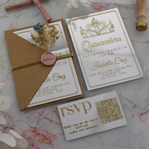 Op de afbeelding: Elegante quinceañera-uitnodigingen met goudfolie letters. De set bevat een kaart met een kroonontwerp, een RSVP-kaart met een QR-code en een uitnodiging met een bruine envelop verzegeld met een roze wasstempel en droogbloemen.