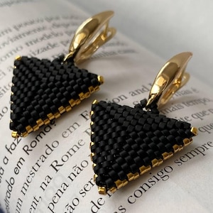 May include: A pair of black beaded triangle earrings with gold-coloured accents. The earrings have a gold-coloured clasp and are displayed on a book page with text.
