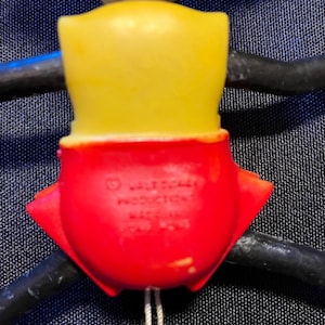 Puede incluir: Una figura de juguete vintage con cabeza amarilla y cuerpo rojo. La figura tiene brazos negros y una cuerda blanca adjunta. El texto "Walt Disney Productions" es visible en el cuerpo rojo.