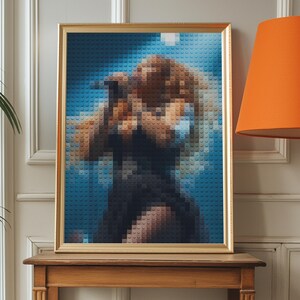 Peut inclure: Une œuvre d'art encadrée représentant un portrait créé avec de petits blocs colorés. L'image a un cadre doré et représente une personne aux cheveux brun clair et une tenue bleu foncé sur un fond bleu. L'œuvre est exposée sur une table en bois.