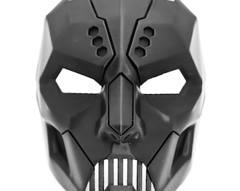 May include: A black, geometric mask with sharp angles and eye openings. The mask features a pronounced nose and a mouth area with vertical lines, resembling teeth. The design is symmetrical and futuristic, with a matte finish.