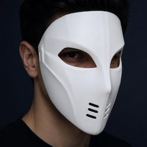 Puede incluir: Una máscara facial blanca y lisa con grandes aberturas angulares para los ojos y una serie de tres ranuras de ventilación horizontales a cada lado de la parte inferior. La máscara presenta un acabado mate limpio y sólido.