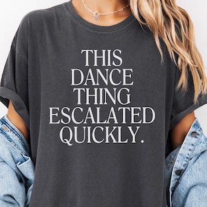May include: Dark gray t-shirt with the text "THIS DANCE THING ESCALATED QUICKLY" in white. The shirt is worn with a light blue denim jacket. The overall style is casual and relaxed.