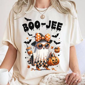 May include: Cream-colored t-shirt with the text "BOO-JEE" above a cartoon ghost wearing sunglasses, a bow, and holding a pumpkin bag and coffee. Black bats and orange pumpkins are scattered around the ghost.