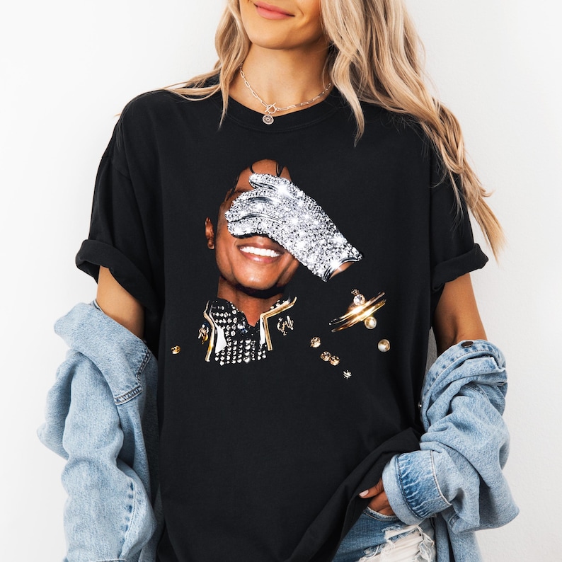 May include: Black t-shirt featuring a graphic of a smiling person with a sparkling glove covering one eye. The design includes gold and silver embellishments. The shirt is worn by a person with long blonde hair.
