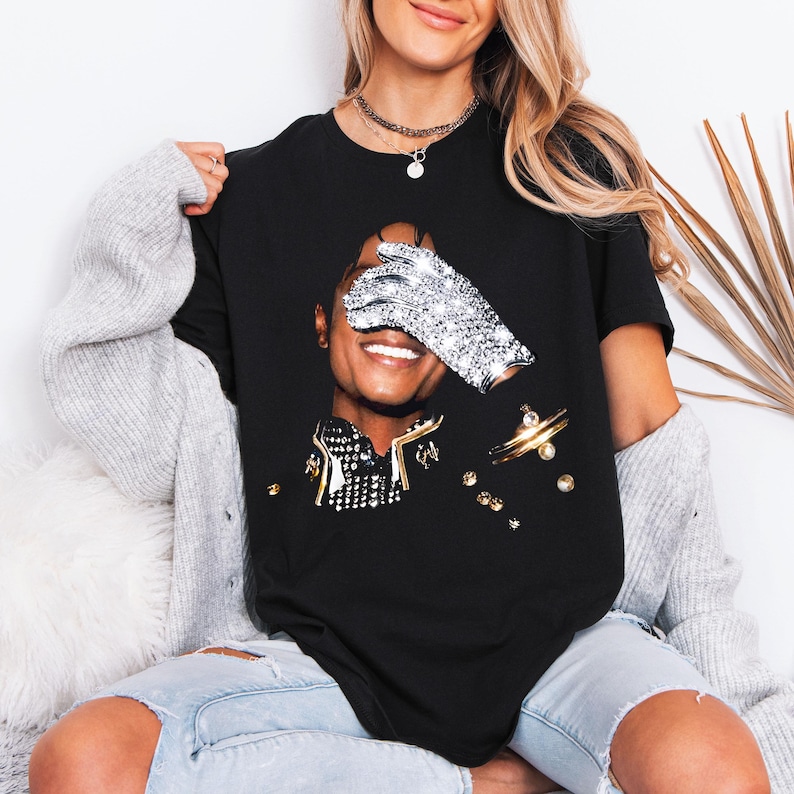 Pode incluir: Camiseta preta com um retrato de uma pessoa sorrindo, um olho coberto por uma luva brilhante. A camiseta tem detalhes dourados e prateados e enfeites brilhantes. O design &eacute; uma homenagem a um &iacute;cone da m&uacute;sica.