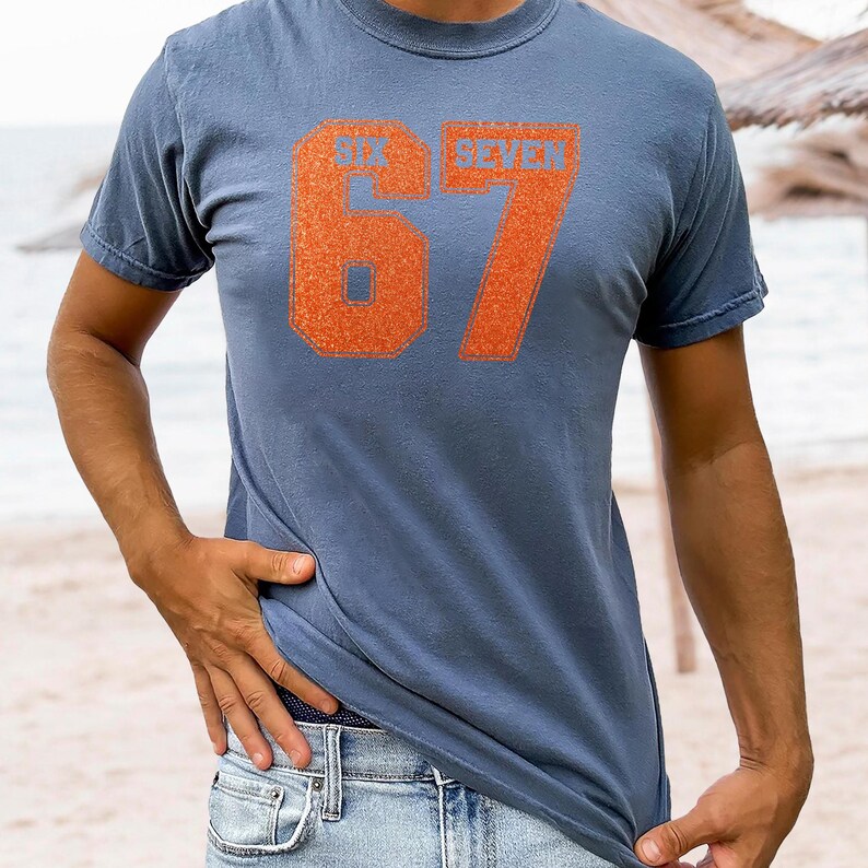 K&ouml;nnte beinhalten: Blaues T-Shirt mit orangefarbenem Aufdruck "SIX SEVEN" und der Zahl "67". Das Shirt hat ein schlichtes Design und besteht wahrscheinlich aus einem weichen Stoff. Der Text ist in einer fetten, blockartigen Schriftart gehalten.
