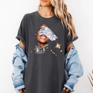 Pode incluir: T-shirt cinza escuro com um gr&aacute;fico de uma pessoa sorrindo, um olho coberto por uma luva de joias. A camisa tem um ajuste folgado e &eacute; combinada com uma jaqueta jeans. O gr&aacute;fico inclui detalhes dourados e prateados.