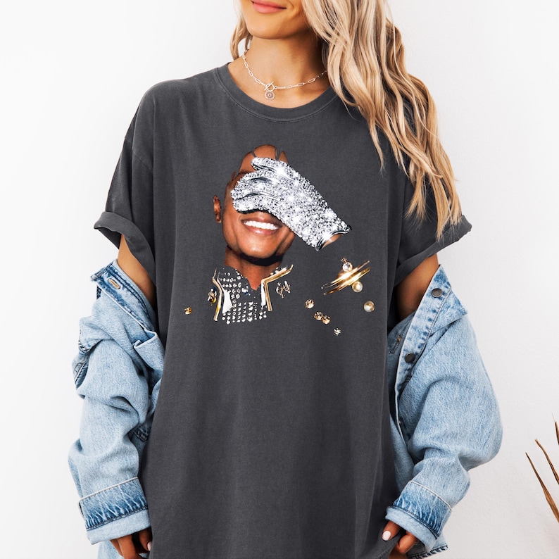 Pode incluir: T-shirt cinza escuro com um gr&aacute;fico de uma pessoa sorrindo, um olho coberto por uma luva de joias. A camisa tem um ajuste folgado e &eacute; combinada com uma jaqueta jeans. O gr&aacute;fico inclui detalhes dourados e prateados.