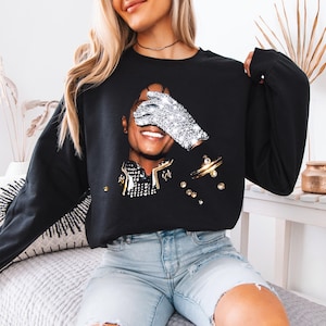 Pode incluir: Camisola preta com um retrato de uma pessoa sorrindo, um olho coberto por uma luva brilhante. O design inclui detalhes dourados e prateados, criando uma est&eacute;tica glamourosa e art&iacute;stica. Esta parte de cima de manga comprida &eacute; um item de moda casual.