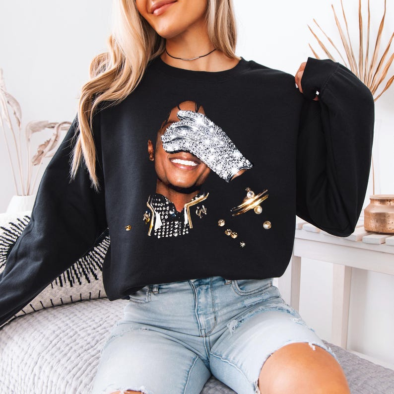 Pode incluir: Camisola preta com um retrato de uma pessoa sorrindo, um olho coberto por uma luva brilhante. O design inclui detalhes dourados e prateados, criando uma est&eacute;tica glamourosa e art&iacute;stica. Esta parte de cima de manga comprida &eacute; um item de moda casual.