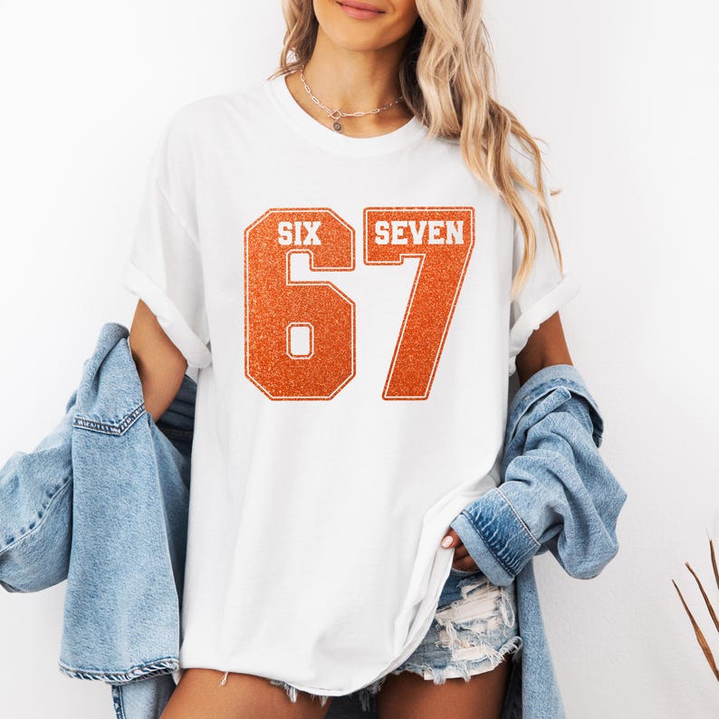 K&ouml;nnte beinhalten: Wei&szlig;es T-Shirt mit der Zahl "67" in orangefarbenem Glitzer-Schriftzug. Das Wort "SIX" steht &uuml;ber der Zahl 6 und das Wort "SEVEN" &uuml;ber der Zahl 7. Das T-Shirt hat hochgekrempelte &Auml;rmel.