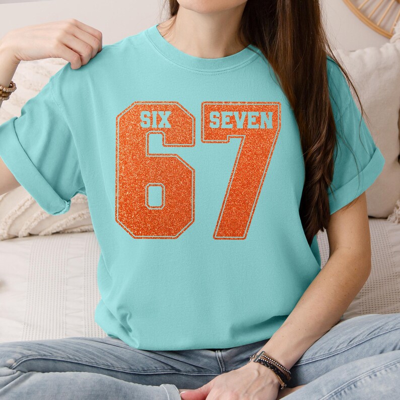 K&ouml;nnte beinhalten: Ein hellblaues T-Shirt mit den Worten "SIX SEVEN" und der Zahl "67" in einer kr&auml;ftigen orangefarbenen Glitzerschrift. Das Shirt hat kurze &Auml;rmel und einen Rundhalsausschnitt.