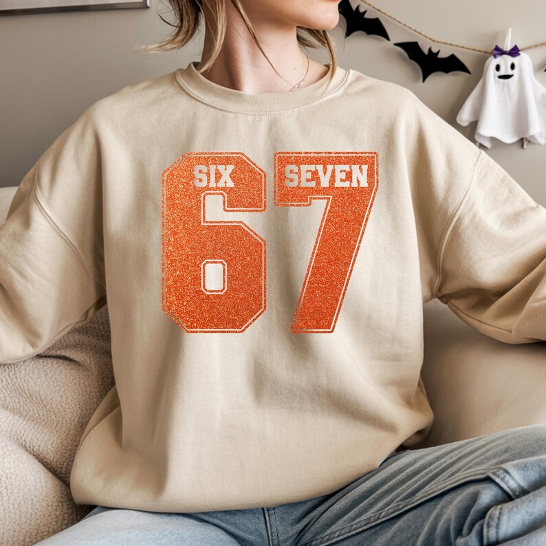K&ouml;nnte beinhalten: Beiges Sweatshirt mit dem Text "SIX SEVEN" und der Zahl "67" in Orange. Die Zahlen haben eine glitzernde Textur. Das Sweatshirt hat eine neutrale Farbe und der Text ist in einer fetten Schriftart im College-Stil gehalten.