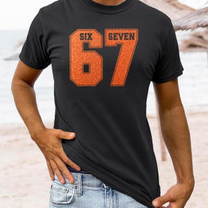 K&ouml;nnte beinhalten: Schwarzes T-Shirt mit den W&ouml;rtern "SIX" und "SEVEN" &uuml;ber der Zahl "67" in einer kr&auml;ftigen orangefarbenen Schrift. Das T-Shirt besteht aus einem weichen Material und eignet sich f&uuml;r Freizeitkleidung.