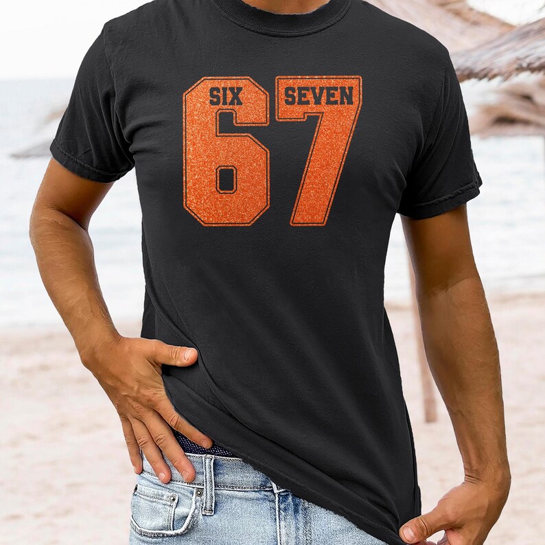 K&ouml;nnte beinhalten: Schwarzes T-Shirt mit den W&ouml;rtern "SIX" und "SEVEN" &uuml;ber der Zahl "67" in einer kr&auml;ftigen orangefarbenen Schrift. Das T-Shirt besteht aus einem weichen Material und eignet sich f&uuml;r Freizeitkleidung.
