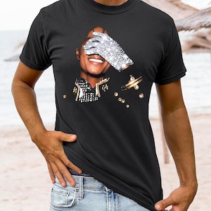 Pode incluir: Camiseta preta com um gr&aacute;fico de uma pessoa usando uma luva de joias sobre o olho. O gr&aacute;fico inclui detalhes dourados e joias espalhadas. A pessoa est&aacute; sorrindo. A camiseta &eacute; usada por uma pessoa em jeans.