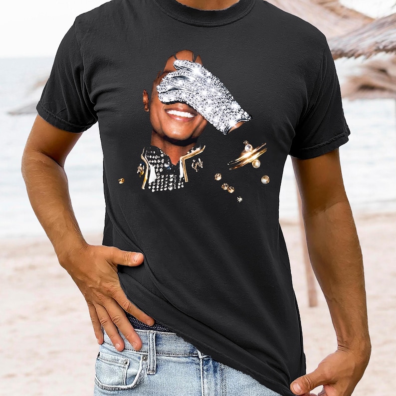 Pode incluir: Camiseta preta com um gr&aacute;fico de uma pessoa usando uma luva de joias sobre o olho. O gr&aacute;fico inclui detalhes dourados e joias espalhadas. A pessoa est&aacute; sorrindo. A camiseta &eacute; usada por uma pessoa em jeans.