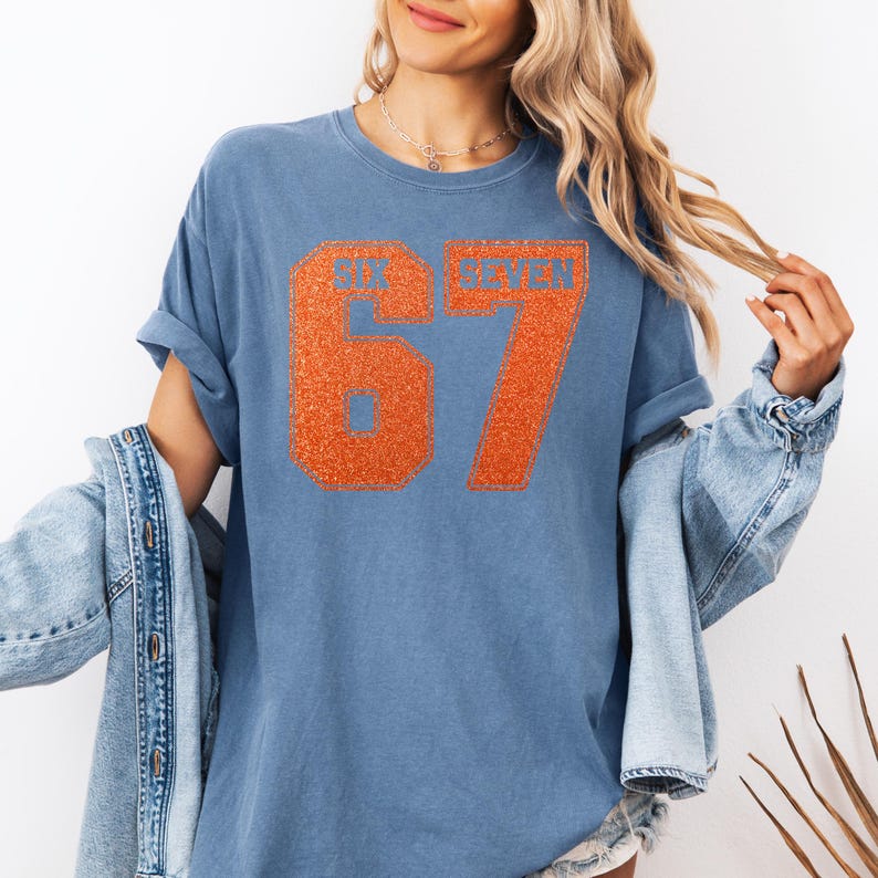 K&ouml;nnte beinhalten: Ein blaues T-Shirt mit dem Text "SIX SEVEN 67" in orangefarbenem Glitzer. Das Shirt hat kurze &Auml;rmel und eine lockere Passform. Eine Jeansjacke ist &uuml;ber den Schultern getragen.