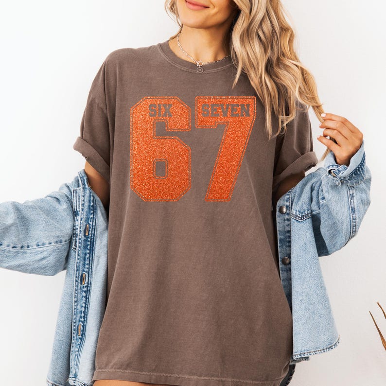 K&ouml;nnte beinhalten: Braunes T-Shirt mit dem Text "SIX SEVEN" und der Zahl "67" in orangefarbenem Glitzer. Das Shirt hat kurze &Auml;rmel und eine lockere Passform. Eine hellblaue Jeansjacke wird dar&uuml;ber getragen.
