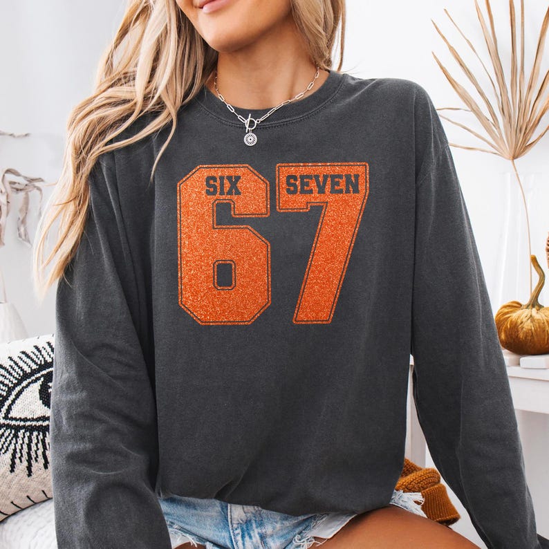 K&ouml;nnte beinhalten: Dunkelgraues Sweatshirt mit der Zahl 67 in orangefarbenem Glitzer, mit den W&ouml;rtern "SIX" und "SEVEN" &uuml;ber den Zahlen. Das Sweatshirt hat einen Rundhalsausschnitt.