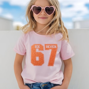 K&ouml;nnte beinhalten: Ein hellrosa T-Shirt mit den Worten "SIX SEVEN" &uuml;ber der gro&szlig;en orangefarbenen Zahl "67". Das T-Shirt wird von einer Person getragen, die eine rosa herzf&ouml;rmige Sonnenbrille tr&auml;gt. Die Person l&auml;chelt.