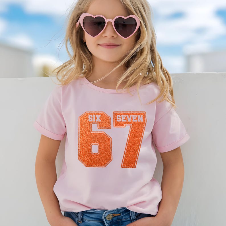 K&ouml;nnte beinhalten: Ein hellrosa T-Shirt mit den Worten "SIX SEVEN" &uuml;ber der gro&szlig;en orangefarbenen Zahl "67". Das T-Shirt wird von einer Person getragen, die eine rosa herzf&ouml;rmige Sonnenbrille tr&auml;gt. Die Person l&auml;chelt.