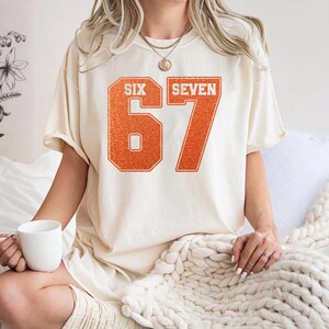 K&ouml;nnte beinhalten: Cremefarbenes T-Shirt mit dem orangefarbenen Text "SIX SEVEN" und der Zahl "67" in einer College-Schriftart. Das Shirt hat eine lockere Passform und kurze &Auml;rmel. Eine Person tr&auml;gt das Shirt und h&auml;lt einen wei&szlig;en Becher.
