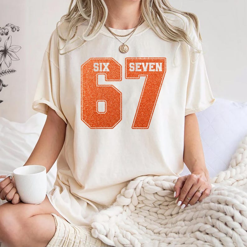 K&ouml;nnte beinhalten: Cremefarbenes T-Shirt mit dem orangefarbenen Text "SIX SEVEN" und der Zahl "67" in einer College-Schriftart. Das Shirt hat eine lockere Passform und kurze &Auml;rmel. Eine Person tr&auml;gt das Shirt und h&auml;lt einen wei&szlig;en Becher.