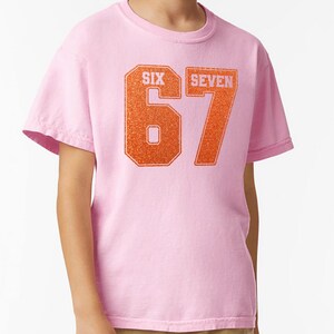 K&ouml;nnte beinhalten: Ein hellrosa T-Shirt mit der Zahl "67" in orangefarbenem Glitzer-Schriftzug. Die W&ouml;rter "SIX" und "SEVEN" stehen &uuml;ber den Zahlen. Das T-Shirt hat kurze &Auml;rmel und einen Rundhalsausschnitt.