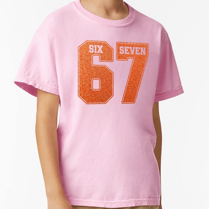 K&ouml;nnte beinhalten: Ein hellrosa T-Shirt mit der Zahl "67" in orangefarbenem Glitzer-Schriftzug. Die W&ouml;rter "SIX" und "SEVEN" stehen &uuml;ber den Zahlen. Das T-Shirt hat kurze &Auml;rmel und einen Rundhalsausschnitt.