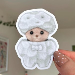 May include: A die-cut sticker featuring a small, light brown bear character wearing a textured, light grey, puffy, segmented suit that covers the head and body, with a small bow tie detail at the neck and a white border outline.
